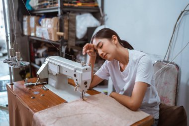 Yorgun kadın terzi perdeyi dikerken terini süpürüyor. Pencere bakım evindeki dikiş makinesini kullanıyor.