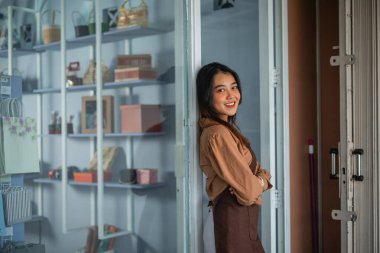 Önlüklü kız ellerini çapraz yapıp gülümsüyor ve hediyelik eşya dükkanında kameraya bakıyor.