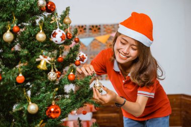 Neşeli bir Noel Baba şapkası takan neşeli bir kadın güzel bir Noel ağacını süslüyor.