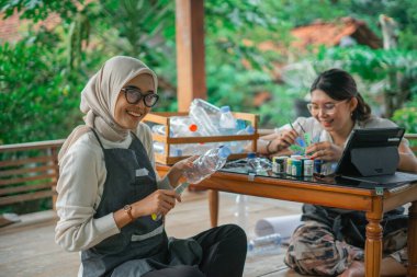 Kafiyeli kadın arkadaşıyla plastik şişe atıklarından el işi yapıyor.