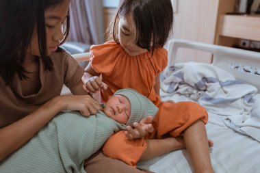 İki kız kardeş nazikçe sarılıp sarmalanmış, değerli ve yeni doğmuş kardeşleriyle etkileşim halindeler.