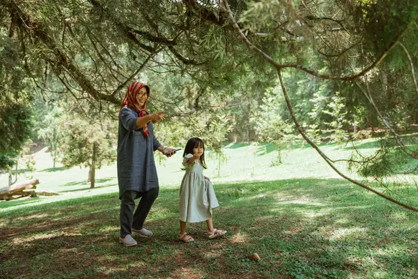 Bir anne ve kızı, uzun ağaçlar ve canlı doğayla çevrili, yemyeşil bir parkta birbirlerine bağlanırlar.