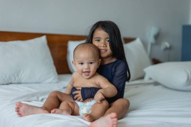 Küçük bir kız sevinçle kucağında küçük bir bebek, yumuşak bir yatakta beşiği kucaklıyor, aralarında sıcak ve sevgi dolu bir sahne yaratıyor.