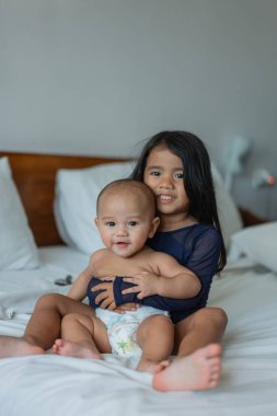 Neşeli bir suratı olan küçük bir kız, bir bebeği nazikçe sıcak bir yatakta tutuyor, sevgi dolu, doyurucu bir sahne yaratıyor.