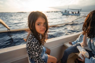 Küçük bir kız, bulutlarla dolu parlak gökyüzünün altındaki engin okyanusta usulca yüzen küçük bir teknede mutlu bir şekilde oturuyor.