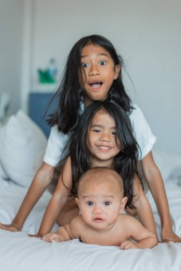 Üç neşeli çocuk, küçük bir bebekle yumuşak bir yatakta neşeli bir atmosferde birlikte gülüp eğleniyorlar.