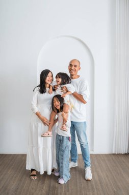 Neşeli bir aile, sade beyaz bir duvarın önünde eğlenceli anları için zemin görevi gören unutulmaz bir resim için mutlu bir şekilde poz veriyor.