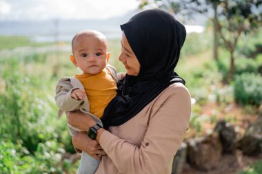 Geleneksel tesettürlü bir kadın, kucağında küçük bir bebeği nazikçe kucaklar ve birlikte şefkatli ve şefkatli bir an yaşar.