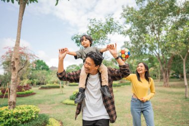 Küçük çocuk, annesi parkta oynarken elinde top tutunca babasının omzunda mutlu oluyor.