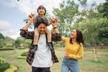 Küçük bir çocuğun babasının omzunda annesiyle parkta top oynarken ki keyfi.