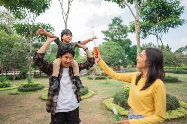 Baba bir çocuğu omzundan tutuyor ve anne parkta oynarken köpük yapıyor.