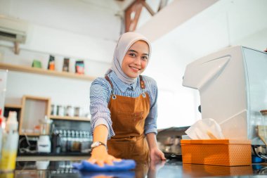 Müşteriler kafeye gelmeden önce masaları temizlerken gülümseyen türbanlı çekici bir kadın.