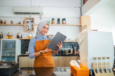 Kafe garsonluğu yapan Asyalı bir kadın kafeteryada tablet kullanarak önlük giyiyor.