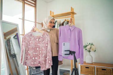 Örtülü kadın bir atölyede cep telefonuyla video kaydederken iki gömlek tutuyor.