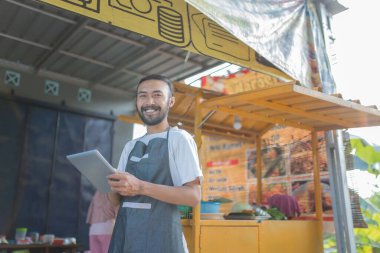 Neşeli bir satıcı, canlı bir açık hava yiyecek standında, müşterileri cezbeden bir tableti ustaca kullanır.
