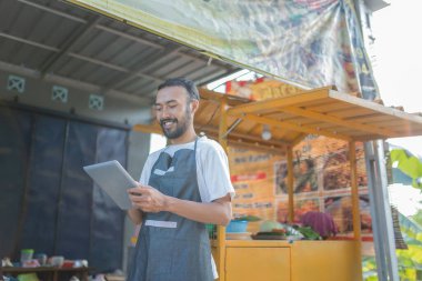 Neşeli bir satıcı, canlı bir açık hava yiyecek standında gururla dururken elinde tablet tutuyor.
