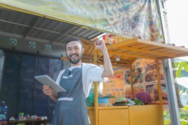 Neşeli bir satıcı, canlı bir sokak yemekhanesinde tablet kullanarak başarılarını kutluyor.
