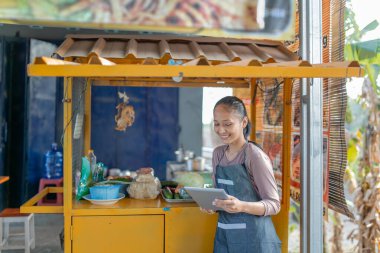 Neşeli bir kadın, canlı bir yemek standında beklerken tabletini mutlulukla kullanıyor.