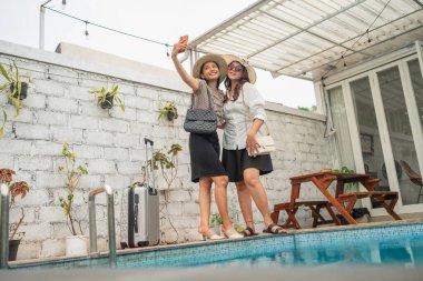 İki arkadaş güneşli, rahat bir tatil atmosferinde havuz başında selfie çekiyorlar.