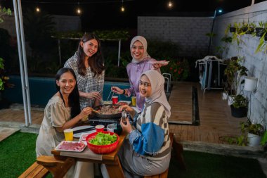 Bir grup arkadaş, parıldayan gece ışıkları altında nefis bir açık hava barbekü yemeğinin tadını çıkarıyorlar.