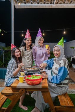 Dostlar dışarıda canlı bir barbekü kutlaması yaparlar, sıcacık renkli parti şapkaları giyerler.