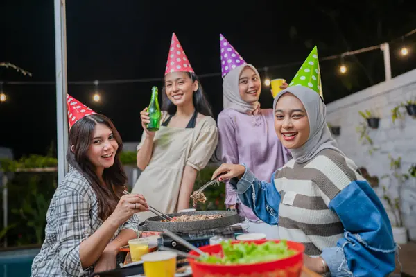 Bir grup arkadaş, gece barbekü, içki ve renkli parti şapkalarıyla birlikte neşeyle kutluyorlar.