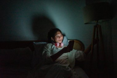 Bir insan rahat ve loş bir yatak odasında cep telefonuyla eğlenirken bulundu.