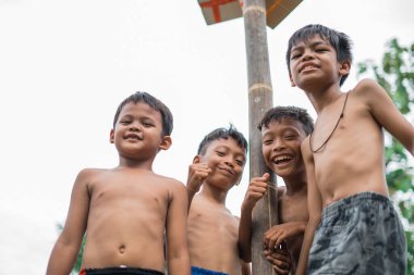 Çocuklar betel ceviz tırmanma yarışmasının direğinin altında dururken kameraya gülümsüyor.