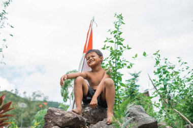 Sırıtkan çocuk, elinde sopayla kayanın üzerinde oturuyor ve arkasında Endonezya bayrağı var.