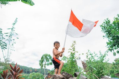 Arka planda doğa gururla Endonezya bayrağını sallayan üstsüz çocuk.