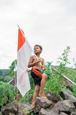 Bağımsızlık kutlamaları sırasında bir kayanın üzerinde duran Endonezya bayrağı taşıyan üstsüz Cava 'lı çocuk.