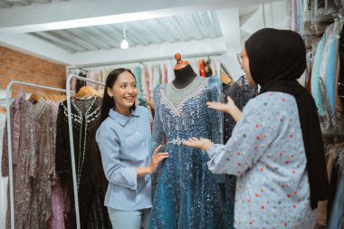 İki kadın, modaya uygun bir butikte şık bir mavi elbiseye bakarken heyecanlı bir şekilde sohbet ediyorlar.