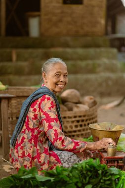 Yaşlı bir kadın, geleneksel pazar ortamında titizlikle taze malzemeler hazırlarken neşeyle gülümser.