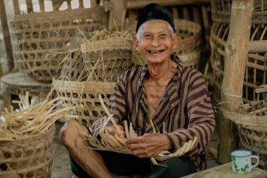 Yaşlı, neşeli bir zanaatkar kırsal bir atölyede bambu sepetleri yapıyor ve geleneksel zanaat becerilerini sergiliyor..