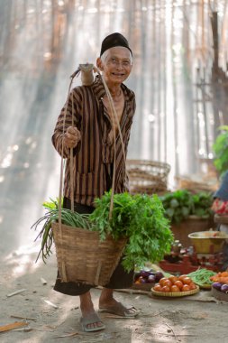 Neşeli yaşlı bir çiftçi, güneşli pazar ortamında bir dizi taze ve canlı ürün sergiliyor.