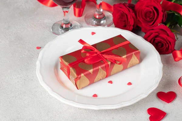 Gift in wrapping paper on white plate, red roses and two glasses for wine on light table, concept for Valentines Day