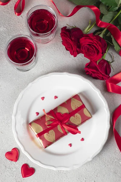 Gift in wrapping paper on white plate, red roses and two glasses wine, concept for Valentines Day, Top view