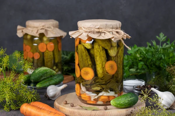 Homemade pickled cucumbers with carrots, garlic, bay leaves and dill in two jars on wooden board