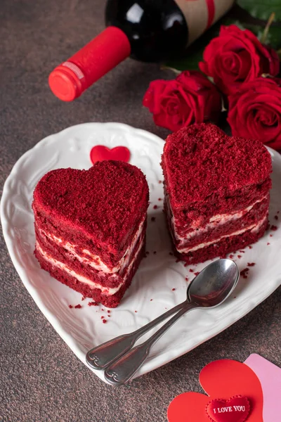 Cakes Red velvet in the shape of hearts on white plate, roses and wine for Valentines Day on brown background