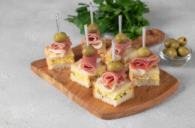Canape with jamon and olives on toasted white bread with cheese on wooden board on gray table