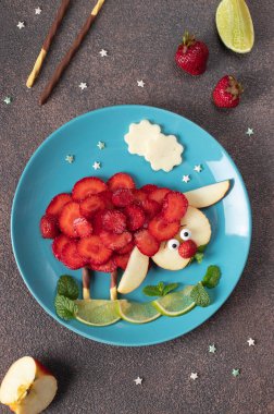 Çocuklar için eğlenceli yemek fikri - çilekli, elmalı ve limonlu koyun hayvan kahverengi arka planda mavi tabağa serilmiş, yukarıdan görünüm