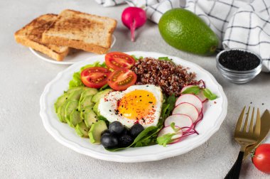 Sağlıklı romantik kahvaltı. Avokado, ıspanak, kinoa, vişneli domates, turp ve beyaz tabakta zeytinle servis edilen kalp şeklinde sahanda yumurta.