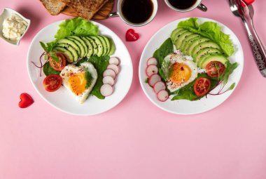 Romantik çift kahvaltısı. İki porsiyon kalp şeklinde kızarmış yumurta. Avokado, ıspanak, vişneli domates ve pembe arka planda turp.