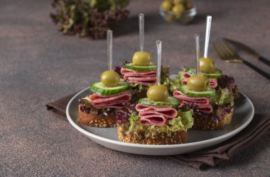 Sosisli, salatalıklı, zeytinli ve marullu kanepe gri bir tabakta beyaz ekmek tostu.