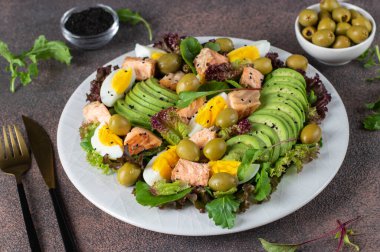 Fırında somon, avokado, yumurta ve zeytinli salata. Kahverengi arka planda beyaz bir tabakta susam tohumları serpiştirilmiş.