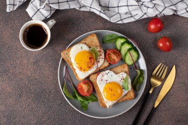 Kalp şeklinde kızarmış yumurta, vişneli domates, salatalık ve kahverengi masada kahvaltı için bir fincan kahve.