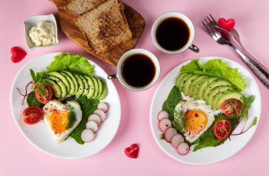 Romantik çift kahvaltısı. İki porsiyon kalp şeklinde kızarmış yumurta. Avokado, ıspanak, vişneli domates ve pembe arka planda turp.