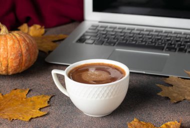 Bir fincan kahve ve dizüstü bilgisayar, sonbahar yapraklarının yanında ve kahverengi masada balkabağı, evde çalışma kavramı