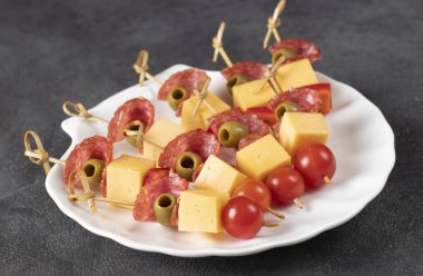 Beyaz tabakta vişneli domates, peynir, sosis ve yeşil zeytin.