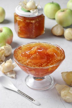 Ev yapımı şeffaf reçel fizsalis ve kasede elmalar ve gri arka planda cam kavanoz.